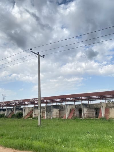 In Rahwol Chom, Nigeria, a weather-beaten farmers market stands under a cloudy sky. The structure, built with concrete and red roofing, features a long, covered walkway supported by a metal frame. Stone stairs lead up to entrances on either side of a central pole. The market is surrounded by lush green grass and power lines crisscross the sky. The overall scene conveys a sense of neglect and disuse.