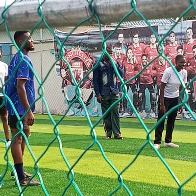 The image depicts an outdoor scene at an artificial turf soccer field in Koko, Nigeria, observed through a green chain-link fence in the foreground. Four individuals are visible on the field. On the left, a young man wearing a blue patterned short-sleeved shirt, black shorts, dark socks, and soccer cleats stands facing left with his head tilted upward. To his immediate left, another individual in a white t-shirt is partially visible. In the middle-right, a man wearing a dark blue denim jacket, dark green trousers with a red side stripe, and brown sandals stands facing slightly right, with his right hand near his temple. On the far right, a young woman wearing a white short-sleeved t-shirt, black trousers, and white shoes stands smiling, facing towards the right. The background features a large banner displaying the Manchester United club crest on the left and a lineup of multiple Manchester United players in red jerseys on the right. A light blue building is partially visible behind the banner on the left side of the frame.