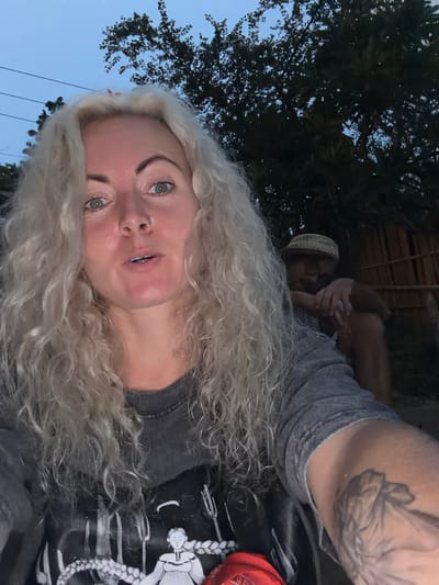 In this selfie, a woman with long, curly blonde hair and blue eyes is in the foreground, facing the camera and speaking. She is wearing a grey t-shirt with a black graphic print and has a nose ring. Her left arm, visible on the right side of the frame, has a tattoo of a feather. Behind her, slightly out of focus, a person wearing a straw hat and glasses is seated, leaning on their knees. They are positioned outdoors, under a tree with dark foliage. The background suggests a natural or rustic setting, possibly a garden or outdoor seating area, with a hint of a bamboo-like structure to the right. The lighting indicates it is dusk or early evening. While the image is taken outdoors, specific details pinpointing a location within Luang Prabang, Laos, are not discernible from the visual information provided.