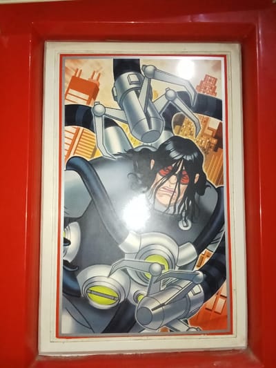 A vertically oriented photograph captures a framed, comic book or anime-style illustration. The illustration depicts a male character with long, dark, wavy hair and red rectangular sunglasses, his mouth open in an apparent strained or determined expression. He is integrated into a prominent, dark grey robotic apparatus featuring multiple silver-colored, articulated mechanical arms extending from his back. Each arm is composed of cylindrical segments and terminates in a three-pronged claw. The apparatus also incorporates circular elements with yellow luminous centers on its front. The character wears a dark, form-fitting garment with black and grey striped sections beneath the robotic components. The background of the illustration shows an urban landscape comprised of orange and reddish-brown high-rise buildings. The illustration is bordered by a white inner frame, which is then enclosed by a wider, glossy, bright red outer frame. A significant glare is visible across the reflective surface covering the illustration, obscuring parts of the character's face and upper body. The specific location within the city of Inxuba Yethemba Local Municipality, South Africa, cannot be determined from the visual content of the image.