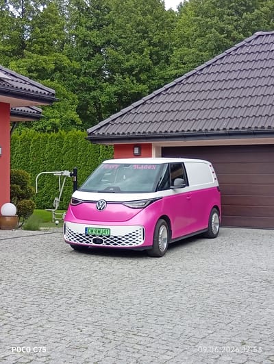 A pink and white Volkswagen ID. Buzz Cargo van is parked on a cobblestone driveway in front of a house in Wilczyce, Poland. The van has a white roof and white front grille with a Volkswagen logo in the center. The license plate reads "DW-8WU41". Pink lettering on the windshield says "PUSSY WAGON". The house has a red exterior and a dark tiled roof. A brown garage door is visible. To the left of the van, there is a white metal exercise machine and a spherical white lamp on a stand. A trimmed green hedge and trees are in the background. The timestamp "09.06.2026 12:53" is visible in the bottom right corner.