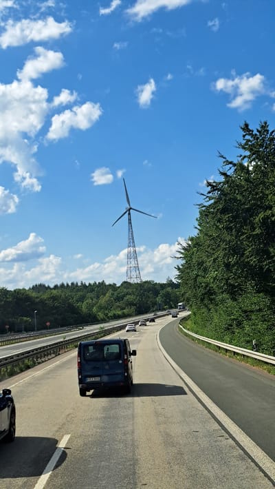 The image captures a view from a vehicle traveling on a multi-lane highway, likely in or near Freudenberg, Germany, given the presence of a prominent wind turbine characteristic of the region. The highway curves gently to the right, bordered by dense green trees on the right and a median with guardrails and more trees on the left. Several cars and a truck are visible on the highway, all moving in the same direction. A dark blue minivan is in the foreground, in the right lane. In the background, a tall wind turbine with three blades stands against a bright blue sky with scattered white clouds. The overall scene depicts a typical highway landscape with a focus on transportation and renewable energy infrastructure.
