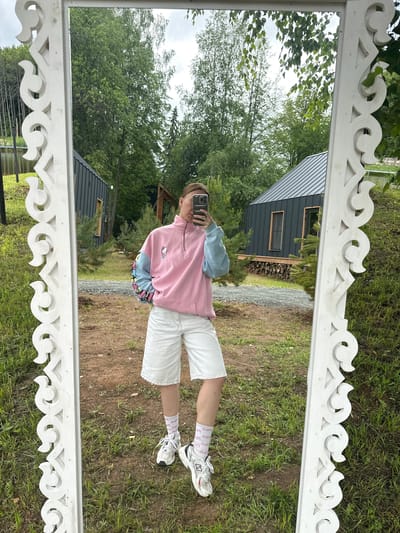 A person stands in front of a mirror outdoors, taking a selfie. They are wearing a pink and blue half-zip sweatshirt with a Hello Kitty graphic on the front, white denim shorts, white socks with a pink pattern, and white athletic shoes. The mirror has an ornate white frame with carved details. The background is a natural setting with green grass, trees, and two small dark-colored buildings with metal roofs. The ground in front of the mirror is a mixture of dirt and gravel. The image is likely taken in Novaya Vozhoika, Russia, given the surrounding natural environment and architectural style of the buildings, which are characteristic of rural Russian dachas.