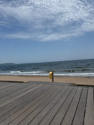 The image depicts a seaside scene in Mariners Bay, Ireland. The foreground is dominated by a weathered wooden boardwalk, its planks running diagonally from the bottom right to the top left of the frame. The boardwalk is bordered by a sandy beach. Beyond the beach, the ocean stretches out, with waves gently rolling onto the shore. The water appears to be a deep, textured blue-grey. The sky above is a pale blue, with scattered, wispy white clouds.

A small, yellow object, possibly a trash receptacle or a small information box, is mounted on a dark post, situated at the edge of the beach where it meets the boardwalk. The scene appears to be set during the daytime, with the lighting suggesting it could be morning or late afternoon. The weather seems mild and possibly a bit breezy, given the texture of the clouds and the movement implied in the waves. There is no visible text in the image. The overall mood is serene and natural, highlighting the meeting point of man-made structure and the coastal environment.