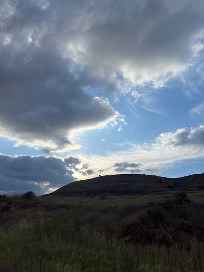 The image shows a view of the sky and a hillside in Ait Mazigh, Morocco. The sky is partly cloudy with both dark and bright clouds. Some areas of blue sky are visible. The hillside is covered in grass and appears to be sparsely vegetated with some rocks visible.