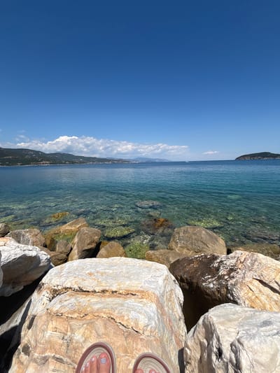 The image depicts a coastal scene in Νέα Ηρακλείτσα, Greece. In the foreground, large rocks of varying sizes and colors, primarily beige and brown, are visible. The lower part of the frame shows a person's feet wearing sandals, indicating they are sitting or resting on the rocks. Beyond the rocks, clear turquoise water stretches out, revealing the seabed beneath. In the middle ground, a coastline with a mountainous backdrop is visible, dotted with buildings. A small island is situated in the distance to the right. The sky is a vibrant blue with scattered white clouds, suggesting a clear, sunny day. The overall scene conveys a tranquil beach or seaside environment.