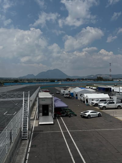 The image depicts an outdoor scene at what appears to be a racetrack in Amozoc de Mota, Mexico, given the presence of mountains in the background. The view is from an elevated position, looking down onto a pit area with several vehicles, including white vans, a white car, and race cars. Structures like tents and metal scaffolding are visible, along with a large transport truck marked with "RAM" logos. The ground is asphalt marked with painted lines. The sky is blue with scattered clouds.