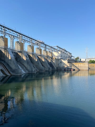 A wide shot captures the impressive scale of the N. B. N. dam, a concrete structure spanning across a tranquil body of water. The dam's angled spillways, cast in shadow by the morning sun, descend towards the water's surface, reflecting the stern geometry of the architecture. A network of overhead metal walkways and supporting structures extends across the top of the dam, hinting at the complex machinery and operations within. The clear blue sky above provides a stark contrast to the massive, man-made edifice. In the foreground, the water is calm, with subtle ripples distorting the reflections of the dam. A small, flat-bottomed boat is moored to a platform near the dam's central section, suggesting human presence and activity in this industrial landscape. The overall scene is one of immense power and engineering prowess, set against the natural backdrop of a clear sky and calm water. The specific location of N. B. N. dam in Ustia, Moldova is indicated.