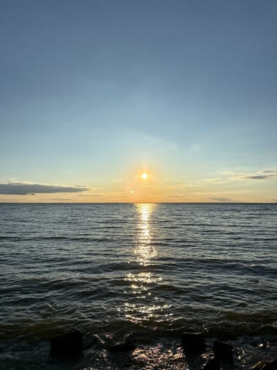 The image displays a waterfront scene at sunset or sunrise over the Kyiv Reservoir in Lebedivka, Ukraine. A bright, circular sun is positioned low above the horizon, slightly right of the frame's center. A prominent, shimmering path of sunlight extends from the sun directly across the dark, rippled water towards the viewer. The sky transitions from light blue at the zenith to a warm yellow-orange hue near the horizon, with scattered, elongated clouds visible on the left and right. In the immediate foreground, dark, irregular objects, consistent with rocks or submerged debris, are visible at the water's edge. No individuals or identifiable man-made structures are present in the scene.