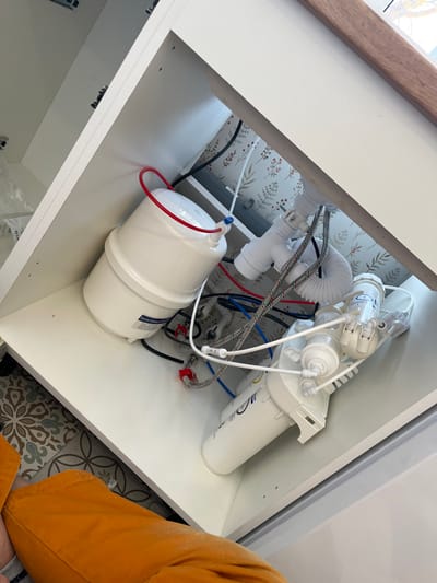 The image displays the interior of a white cabinet, likely beneath a sink, containing a water filtration system and plumbing components. A white, cylindrical pressure tank for reverse osmosis is positioned on the left, connected by a red tube at its top. To the right, multiple white water filter housings are interconnected by blue, red, white, and black plastic tubing and various fittings. A white plastic P-trap and a flexible white corrugated drain hose are visible in the upper right, along with two braided stainless steel water supply lines connected to valves. The cabinet's rear wall is covered with light wallpaper featuring a repeating botanical pattern of green leaves and small red elements. A wooden countertop edge is partially visible at the top right. In the lower-left quadrant, the legs of an individual wearing orange pants are visible. The floor outside the cabinet features patterned tiles. The location of Нарынка, Russia, cannot be confirmed from the visual information present in the image.