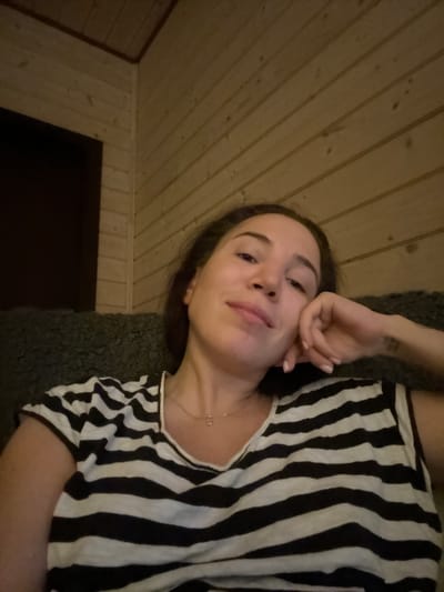 A woman is taking a selfie in a room with wood-paneled walls. She is wearing a black and white striped shirt and a necklace with a small pendant. Her head is resting on her hand. The room appears to be dimly lit. There is no discernible information in the image to indicate the location is the city of Stupino, Russia.