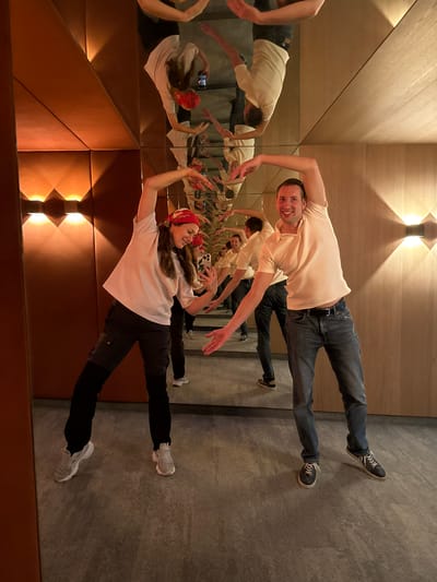 Two people, a woman and a man, are posing with their arms outstretched in a mirrored room. The woman, on the left, wears a white t-shirt, black pants, and a red bandana. The man, on the right, wears a light-colored polo shirt and blue jeans. The room features wooden paneled walls and ceiling, with modern wall-mounted light fixtures. The mirrored surfaces create numerous reflections of the subjects and the room, making it difficult to determine the exact dimensions of the space. The floor is a textured gray surface. It is not possible to determine the location in Mayrhofen, Austria from the image alone.