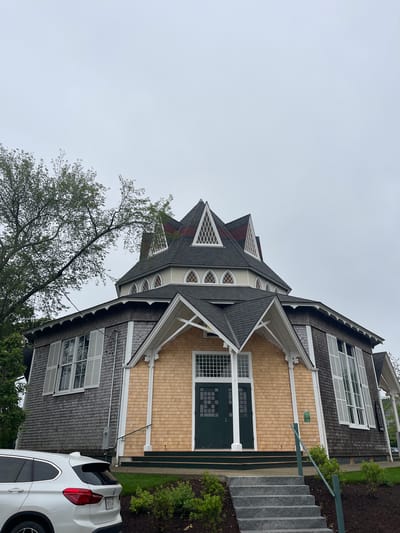 The image shows the Flying Horses Carousel in Oak Bluffs, Massachusetts, United States of America, on an overcast day. The carousel building is octagonal and features a distinctive multi-tiered roof with pointed gables and arched windows. The exterior walls are covered in cedar shingles, with a section of light-colored cedar shingles on the front facade. A white BMW SUV is parked in the foreground to the left, partially obscuring the view of the building's base. A set of stone steps leads up to the entrance of the carousel, which has dark green double doors. Green railings line the steps and the walkway. Lush green bushes and manicured grass surround the building and steps. A large tree with green foliage is visible to the left of the building. The sky is a uniform light gray.