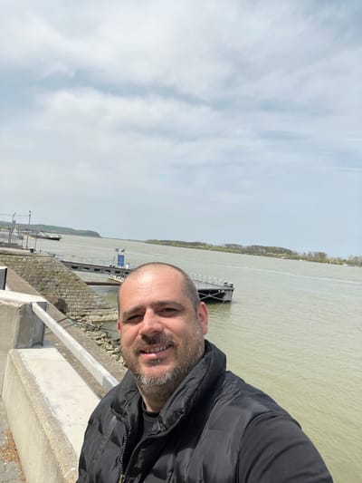 A selfie of a smiling man with a shaved head and a dark jacket. Behind him is a river with a platform dock. The background also includes trees along the far side of the river. The sky is overcast. This image may be taken at the Nikopol, Bulgaria riverfront, based on the river and dock features.