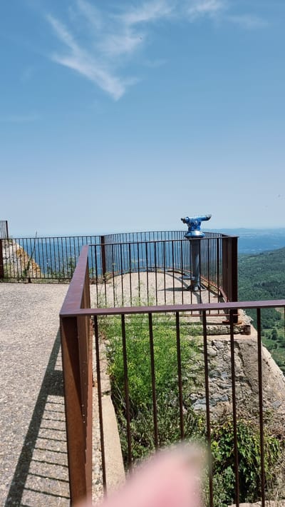 This is a full, horizontal, outdoor shot of a scenic viewpoint in Susqueda, Spain. In the foreground, a rusted metal railing runs along the left and bottom of the frame, casting a sharp shadow on a textured concrete path. To the right of the railing, lush green vegetation grows around a rocky incline. In the midground, another rusted metal railing encloses a circular viewing platform paved with gravel. A blue, coin-operated telescope is centrally positioned on the platform, facing the distant landscape. The background features a vast expanse of blue sky with scattered wispy white clouds. Below the sky, a hazy blue horizon stretches across the image, revealing a sprawling green landscape of rolling hills and distant villages. The scene appears to be captured on a clear, sunny day.