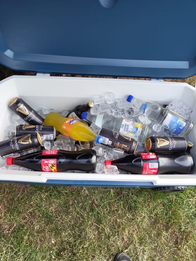 The image shows a blue and white cooler filled with ice and various beverages. The cooler appears to be outdoors on a grassy surface, based on the visible background. Inside the cooler, there are bottles of water (including one labeled "STEINER"), glass bottles of "SCHORLE WASSER," a yellow Fanta bottle, bottles of Bitburger beer, and Coca-Cola bottles. Additionally, there are several cans of Guinness Draught stout. The cans and bottles are surrounded by ice. A portion of a shoe is visible on the right side of the image. The image contains the words "Schmelz, Germany" on one of the labels of the bottled water.