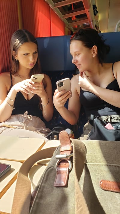 Two young women are seated next to each other, possibly on a train, looking at their smartphones. The woman on the left, with long brown hair, wears a black tank top and light-colored pants. She holds a gold-colored smartphone in her left hand and is looking at it with a serious expression. She has several bracelets on her left wrist and rings on her fingers. The woman on the right, with dark hair pulled back, wears a black tank top and is looking down at her phone, which is also gold-colored. She has earrings in her ears and her right hand is near her chin. Between them, a brown bag with leather accents is visible in the foreground, partially obscuring a table. Behind them, the orange curtains of what appears to be a train carriage are illuminated by sunlight. The environment suggests they are indoors, possibly traveling. The context provided about Ladce, Slovakia, is not visually represented in the image itself.