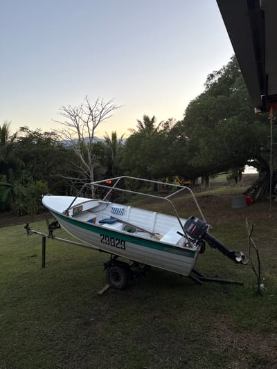 A white and green aluminum fishing boat, bearing the registration number "29829" in black, is situated on a single-axle trailer on a grassy lawn in Dumbéa, France. A black Yamaha 30hp outboard motor is affixed to the boat's transom, with its propeller facing right. The boat's interior contains an oar, a blue scoop, and other small items. A metal frame structure, possibly for a canopy, is installed over the boat. The trailer's front wheel rests on a wooden block. The surrounding environment consists of lush green tropical vegetation, including palm trees, and a prominent bare deciduous tree in the midground. Distant mountains are visible beneath a light blue sky transitioning to orange-yellow at the horizon, indicative of sunrise or sunset. A large, densely foliated tree occupies the right side of the image, partially obscuring a yellow excavator and a glimpse of a body of water or shoreline. A dark architectural overhang, presumably from a building, is visible in the upper right, from which a rope descends. A red bucket and stacked wood are present near the large tree. No individuals are present in the scene.