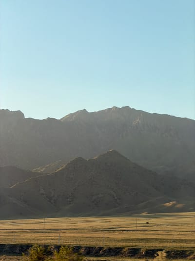 The image shows a mountain range adjacent to a flat expanse of land. The sky is clear and light blue. The mountains are a darker shade of gray and appear rugged. To the right of the mountains is a flat, grassy area with a fence line. Utility poles run alongside the fence. The location is likely in the region of Chüy, Kyrgyzstan, given the prompt.