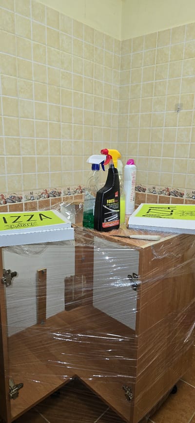The image shows a wooden cabinet wrapped in clear plastic sheeting. On top of the cabinet are several cleaning supplies, including spray bottles and a white container with a pink lid. To the left of the cleaning supplies is a bright green box with the word "PIZZA" in large black letters. The cabinet is placed against a wall tiled with small, beige square tiles. The floor appears to be tiled as well, with larger, brown tiles. A decorative border of smaller, more colorful tiles runs along the bottom of the tiled wall. The overall impression is of a domestic or commercial storage area, possibly a kitchen or utility room. The specific location in Chișinău, Moldova, is not discernible from the image.