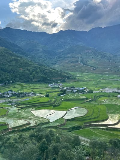 This elevated panoramic image captures a wide mountainous valley in Tú Lệ, Vietnam. The upper portion of the frame features a sky with scattered, primarily cumulus clouds; sunlight penetrates the clouds on the left, casting bright illumination, while clouds on the right appear darker and denser.

Multiple layers of mountains extend across the background, displaying undulating profiles and slopes covered in dense green foliage. Agricultural terracing is visible on many of these mountain sides, receding into the distance.

The mid-ground and foreground are characterized by an extensive network of rice terraces. These terraces exhibit variations in color: some are a vibrant, uniform green, indicating actively growing crops, while others are filled with water, reflecting the sky as bright, silvery-white patches. This creates a mosaic pattern across the valley floor and lower slopes.

Within the valley, several clusters of human habitation are present, consisting of small structures with dark-colored roofs. These settlements are interspersed among the rice fields. A winding, light-colored riverbed or shallow stream is discernible, traversing the valley floor and passing near the human settlements and terraced fields. Narrow, unpaved roads or paths can be observed winding through the terraced landscape, connecting different areas.

The immediate foreground is occupied by the dense canopy of green trees, suggesting the vantage point is from a higher elevation looking down into the valley. No human figures or discernible activities are visible in the scene.