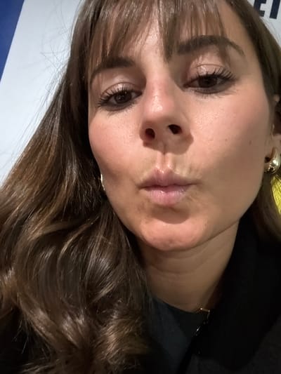 A close-up, angled portrait features a woman with her head tilted, looking towards the viewer. She has long, wavy brown hair with lighter highlights, styled with bangs that frame her forehead. Her dark eyes are open, with visible mascara on her lashes, and she maintains a neutral, calm expression. Her lips are closed, appearing glossy and a natural light pink. Her skin is clear, with a fair to medium tone.
She is wearing a chunky, gold-toned hoop earring in her visible ear, and a thin gold-toned chain or necklace is partially visible around her neck. The background is divided into a dark, shadowed area on one side, likely dark clothing or a backdrop, and a lighter white or light blue surface on the other, against which her head rests.
The woman is posing for the photograph. The lighting is soft and even, without strong shadows, making the time of day or specific weather conditions undeterminable. While the image does not provide explicit geographical markers, the context given is L'Étrat, France. Faint, indistinct blue text, possibly beginning with "L'E", is partially visible on the lighter background surface, obscured by her hair.