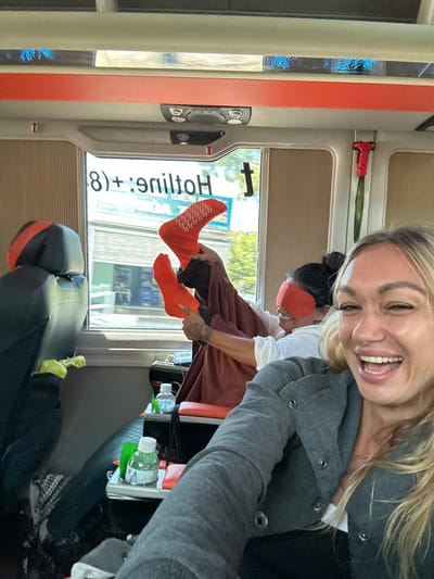 The image shows two women inside what appears to be a bus or van. One woman in the foreground, with blonde hair, is taking a selfie and smiling broadly, her mouth open. She is wearing a grey jacket over a white top. Behind her, another woman with dark hair is wearing a red blindfold and holding up a pair of orange socks with white patterns. She is also smiling. The interior of the vehicle has black leather seats with red accents and brown ribbed panels on the walls. Outside the window, there is a blurred view of buildings and trees, suggesting an urban or suburban setting. There are two bottles of drinks on a small table between the seats. The text "8) + 9niltoH" is visible on the window, which is likely a reversed or mirrored reflection of signage. This text does not directly relate to Hòa Thắng, Vietnam, as it appears to be English words. The overall scene depicts a lighthearted, informal moment.