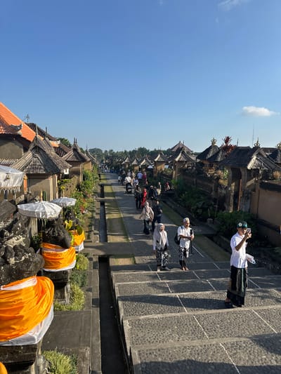This image captures a sunlit scene along a stone pathway lined with traditional Balinese buildings in Bangli, Indonesia. The architecture features tiled roofs with intricate carvings and ornate golden finials. Several people are walking along the pathway, some dressed in traditional attire, including sarongs and head coverings. One individual in the foreground on the right is holding up a mobile phone, appearing to take a photograph. White umbrellas adorned with fringe are positioned on stone structures to the left, draped with bright orange fabric. Lush greenery and tropical plants are visible throughout the scene. The sky is a clear, bright blue with a few wispy clouds. The pathway appears to be a set of stairs made of dark cobblestones, with lighter lines marking the edges of the steps. A narrow channel, possibly for water drainage, runs alongside the pathway. The overall impression is of a cultural site or village, bathed in the warm light of day.