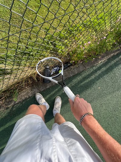 The image displays a first-person, downward-angled perspective of an individual standing on a green textured tennis court surface at a tennis facility in the city of Cherry Burton, United Kingdom. The individual wears white athletic shorts, white socks, and light-colored athletic shoes. Their right hand, adorned with a silver ring on one finger and a metal curb-link bracelet on the wrist, tightly grips the white handle of a white and black Head brand tennis racket. Positioned against a chain-link fence, beyond a concrete curb and a narrow grassy verge to the left, are a black water bottle and a blue and yellow Dunlop-branded can of tennis balls, with at least one yellow tennis ball visible through the racket's strings. The grassy verge contains several small plants exhibiting purple blossoms. The scene is illuminated by direct sunlight, casting shadows from the fence onto the court.
