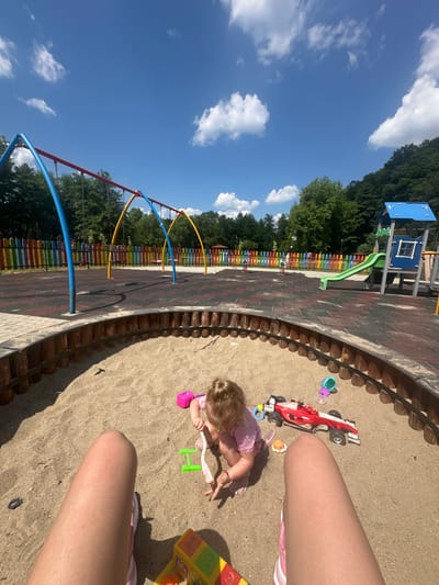 This is a medium shot of a child playing in a sandbox at a playground. The child is a toddler with blonde hair, wearing a pink shirt and shorts. They are surrounded by toys, including a red and white race car, a pink spade, and a green rake. The sandbox is circular and made of wood. In the background, there is a swing set, a slide, and a colorful fence. The playground is located in a park with trees and a blue sky with white clouds. The location is the playground in the city of Donchovci, Bulgaria.