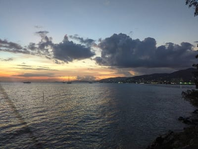 This image captures a twilight scene over the Gulf of Paria, with the city of Port of Spain, Trinidad and Tobago, illuminated along the distant right coastline, backed by the dark silhouette of the Northern Range mountains.
The sky exhibits a gradient from light blue-grey overhead to vivid orange, yellow, and red hues at the western horizon, indicating sunset. Scattered dark grey cumulus clouds are present, with denser formations obscuring portions of the mountains in the background.
The foreground consists of dark, rippling water. A single slender, dark pole-like object protrudes vertically from the water near the center-left. Along the far right edge of the frame, a dark, rocky shoreline with dense, dark green foliage is partially visible.
Multiple vessels are observed on the water. Towards the left horizon, a larger motor vessel or cargo ship is discernible, along with a smaller utility boat and several other indistinct vessels in the mid-distance. Two sailboats with visible masts are positioned centrally on the water, and another smaller boat is further to the right. All visible vessels appear stationary.
The coastline of Port of Spain spans the mid-ground, characterized by numerous artificial lights from buildings and streetlamps. The dark, undulating forms of the Northern Range mountains rise directly behind the city, silhouetted against the twilight sky. No people are visible. No explicit actions or interactions are depicted; the scene is static.