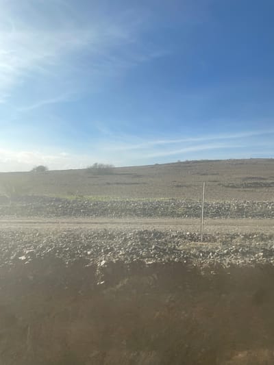 A landscape under a blue sky with wispy clouds. A dirt road runs across a barren field littered with rocks and gravel. A metal post stands in the middle of the field. The terrain appears dry and desolate, transitioning into a darker, shadowed area. Sparse vegetation dots the landscape, hinting at resilient life in the harsh environment.
