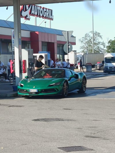 The image depicts a green Ferrari SF90 Stradale parked at a gas station under an "AUTOGRILL" sign. The Autogrill appears to be located in Tortona, Italy, as suggested by the vehicle's license plate, "HB-571SZ," which indicates Italian registration. A young man, possibly the owner, is standing by the car, appearing to clean or adjust something on its front. Several other individuals are visible in the background near the Autogrill building and on the side, with some seemingly interacting or observing. The scene is set in an outdoor, daytime environment with a clear sky. The car is the primary focus, positioned centrally in the frame, while the Autogrill and surrounding elements provide context.