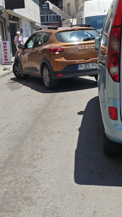 This image shows a street scene in Ataşehir, Turkey. Two cars are parked close together, with the rear of a light blue car partially visible at the top, and a brown Dacia Sandero occupying the majority of the frame. The Dacia has a license plate that reads "34 GPO 162" and the word "DACIA" is visible on the trunk. A person wearing a light-colored hat and shirt is standing next to the driver's side door of the Dacia, appearing to be interacting with the car.

In the background, there are buildings and signs. One sign with white text on a blue background reads "YAPA EMLAK". Another sign with red text on a white background, with an arrow pointing to the right, displays "BERBER". The scene appears to be set during the daytime, with sunlight casting shadows on the asphalt road. The weather seems to be clear and sunny. The overall impression is that of an ordinary day on a city street, with the parked cars and shops suggesting an urban environment.