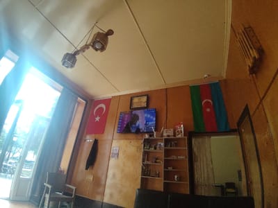 The image depicts the interior of a room, possibly a cafe or lounge, in Khudat, Azerbaijan. A Turkish flag with a white star and crescent on a red background is displayed on the left wall. To its right, an Azerbaijani flag with a crescent and star on a tricolor background of blue, red, and green is visible. A television screen displays an image of a person. Above the television, a framed picture hangs on the wall. A wooden shelf unit is filled with various items, including food packaging and what appear to be other commodities. A light fixture hangs from the ceiling, and a window with sheer curtains allows natural light to enter the room, revealing an outdoor scene with trees and chairs. A chair is positioned in the foreground. The walls are paneled with what appears to be wood.
