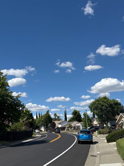The image depicts a suburban street scene in Citrus Heights, California, under a bright blue sky with scattered white clouds. A freshly paved asphalt road with a white dividing line curves to the right, with double yellow lines indicating passing zones. A blue SUV is parked on the right side of the road, facing away from the camera.

On the left side of the road, a dark wooden fence lines the property, with mature trees and some palm trees visible behind it. Several houses with pitched roofs and light-colored exteriors are visible in the background, set back from the street and separated by fences and landscaping. A stop sign is visible on the left side of the street.

On the right side of the road, a sidewalk runs alongside the street, with a neatly trimmed green hedge bordering the properties. More houses and trees are visible in the background on this side as well. Several cars are parked along the street and in driveways. The overall impression is a typical sunny day in a residential neighborhood.