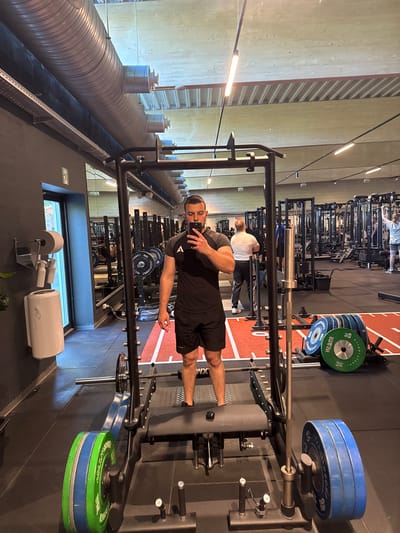 The image depicts a man taking a selfie in a gym setting. He is standing inside a squat rack, wearing black shorts and a dark-colored t-shirt. The squat rack is loaded with weight plates. Behind him, other gym equipment is visible, along with other individuals exercising. The gym has a modern design with exposed ventilation ducts on the ceiling and wooden wall paneling. A window is visible, and cleaning supplies are located in front of it. Based on the presence of the "CROSSFIT ROSKILDE" brand on some of the weight plates, it can be inferred that this image was taken in Roskilde, Denmark.