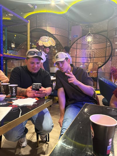 This media file captures two young men indoors, possibly at a restaurant or cafe, given the presence of tables, cups, and decorative lighting. One man, wearing a white baseball cap and a dark t-shirt, is making a peace sign with his fingers while looking at the camera. The other man, also wearing a cap and a black t-shirt, is holding a smartphone and looking towards the camera with a slight smile. They are seated at a table with a black, reflective surface, on which there are several cups and what appears to be a placemat with some text and graphics. The background reveals a modern interior with intricate latticework on the ceiling and walls, and some colorful lighting, suggesting a contemporary establishment. The overall mood appears casual and friendly, with the subjects looking relaxed and engaged with the camera. There is no indication of the time of day or weather conditions. The provided location context is Carretera De San Martín, Spain. Visible text includes "NY" on one of the caps and possibly branding on the cups.