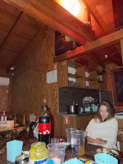 In Zasele, Bulgaria, a dimly lit, rustic room features exposed wooden beams and plywood walls, suggesting an unfinished or unconventional interior. The space appears to be set up for a gathering, with a table in the foreground laden with plastic cups, a jar of yellow substance, a large bottle of Coca-Cola, and what seems to be food remnants. A woman, draped in a white blanket, is seated at the table, engrossed in her phone. In the background, a kitchenette area is visible, equipped with a sink, pots, and shelves stocked with glasses. A fire extinguisher sign is mounted on the wall, and a white air conditioning unit is installed near the ceiling. The overall ambiance is casual and informal.