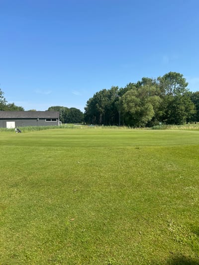 The image shows a well-maintained golf course under a clear blue sky. In the foreground, a vast expanse of neatly mown green grass stretches towards the horizon. To the left, a dark gray building with a sloping tiled roof and white doors is situated. Adjacent to the building, a black golf bag stands upright, with a white golf club leaning against it. Beyond the building and to the right, a line of lush green trees forms a backdrop, with some taller trees interspersed. In the middle ground, another stretch of manicured grass leads to a distant line of vegetation and trees. A green sign is visible on the right side of the image amidst the grass. The overall scene depicts a serene outdoor setting on a sunny day, likely at a golf club in Wassenaar, Netherlands, given the typical landscape of such facilities in the region.