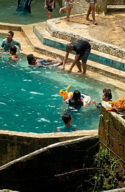 The image captures a scene at a swimming pool in Penungkiren, Indonesia, where several people are enjoying the water. In the foreground, a young boy in a teal t-shirt is visible from the back, looking away from the camera and submerged up to his shoulders in the turquoise water. To his left, an adult male in a black t-shirt and shorts leans over the pool's edge, with his hands near the water's surface, possibly interacting with someone in the pool.
Further into the pool, a group of individuals are present. One person is lying on their back in the water, with another person next to them. Closer to the center, a child is in a yellow and orange inflatable toy shaped like an animal, wearing a pink floatation device. To the right of this child, another person is partially submerged, looking towards the child. In the background, on the pool's edge, several other people are either standing or walking. One person, wearing a dark shirt, is visible from the waist down. Another individual, in a dark dress or shirt, stands with their legs apart, holding a light-colored object. Further back, a person in a black and white patterned garment is also visible, as is someone with light-colored shorts and a white top. The pool has a tiered structure with stone and blue tile accents along the edges, and the surrounding environment appears to be a mix of concrete and natural elements like rocks and vegetation.