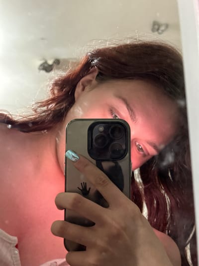 The image is a reflection in a mirror, showing a young person taking a selfie with their phone. The person has reddish-brown hair, which is long and appears to be braided. They are looking at the phone camera, and their eyes are visible, as is part of their forehead and ear. The phone is held in their right hand, and a fingernail with a holographic design is visible. The phone case has a black decal of a handprint. The reflection also shows the person's shoulder and chest area, which appears to be illuminated by pinkish light, possibly from a phone screen or external light source. In the background of the reflection, a blurry image of what looks like a butterfly sticker on the mirror is visible. The overall impression is that of a casual, personal moment captured in a mirrored surface. There is no visible text in the image. The context provided suggests the location is Bishkek, Kyrgyzstan, but there are no specific visual cues in the image to confirm this.