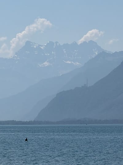 The image depicts a hazy, layered mountain landscape viewed from across a body of water, likely Lake Geneva, in or near Vevey, Switzerland. The foreground is dominated by the blue-grey water, with small ripples suggesting a calm day. A dark, triangular marker or buoy with a black flag is visible protruding from the water on the left side of the frame. In the mid-ground, layers of mountains recede into the distance, their forms softened by atmospheric haze. A distinct structure with a tall, slender chimney, possibly a factory or power plant, is situated on the slope of one of the mountains. The background features more distant, snow-capped peaks, softened by the atmospheric conditions, and a pale blue sky with scattered white clouds. The scene is serene and conveys a sense of vastness and natural beauty, with a touch of industrial presence.