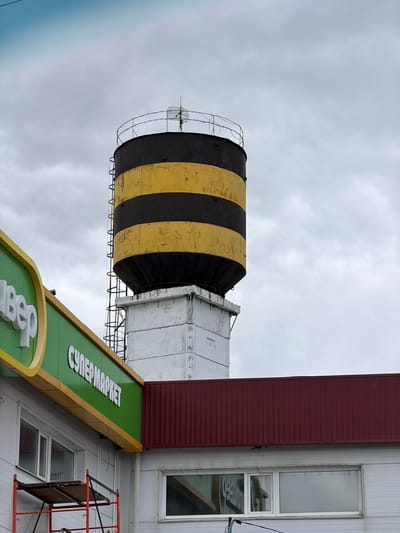The image shows the exterior of a building with a distinctive water tower. The water tower is large and cylindrical, with alternating black and yellow stripes. It appears to be made of metal and is surrounded by a metal cage structure. Attached to the building is a green sign with yellow trim, featuring white text in Russian that reads "СУПЕРМАРКЕТ" (SUPERMARKET). The building itself is clad in white siding, with a dark red section on the upper left. There are several windows visible, some of which are long and narrow. A portion of scaffolding or a similar construction is visible on the right side of the image. The sky is overcast, suggesting a cloudy or possibly rainy day. The scene is shot from a low angle, looking upwards at the water tower and the building. The overall impression is that of an industrial or commercial area, likely in a town or city in Russia. There are no people visible in the image.