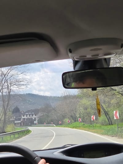 The view from inside a vehicle, driving on a winding road marked with red and white chevron signs. Outside, there are trees and a building with a sign indicating "Adamec Lomnica" visible, likely a hotel or business in the Leshnitsa, Bulgaria area. A driver's hand is visible on the steering wheel.