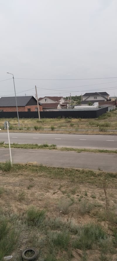This is a medium-shot, outdoor photograph taken in a semi-arid environment, likely Aksu, Kazakhstan, under an overcast sky, suggesting a cloudy day. The scene depicts a suburban or rural residential area bordering a paved road.
In the foreground, dry, sparse vegetation and sandy soil are visible, with a discarded tire lying on the ground to the left. A white pole with a circular sign is also present near the road. The road itself is a two-lane asphalt surface with white lane markings.
Beyond the road, a black metal fence runs horizontally, separating the undeveloped land from a row of houses. These houses appear to be modern, single-family dwellings with various rooflines and architectural styles, some featuring brick exteriors and dark-colored roofs. Electrical poles and wires are visible in the background, crisscrossing the sky.
The overall atmosphere is one of a developing residential area. No people are visible in the frame. The time of day appears to be daytime, given the ambient light, but the cloudy conditions create a muted, diffused light. The location is suggested by the dry, scrubby vegetation and the architecture of the houses, consistent with regions of Kazakhstan. No text is discernible in the image.
