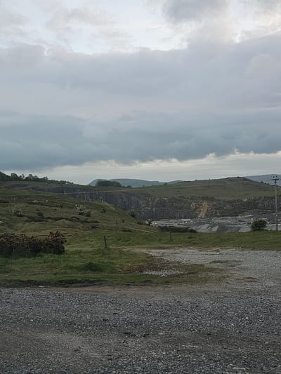 The image depicts a landscape in Halkyn, United Kingdom. It features a quarry with visible rock faces and geological formations. Green vegetation covers the surrounding hills and slopes. A gravel or dirt road runs along the edge of the scene, and utility poles are seen ascending the terrain. The sky is filled with overcast clouds, suggesting potentially gray and muted lighting conditions.