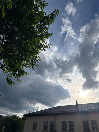 The image depicts a view looking upwards towards the sky from a ground-level perspective. In the foreground, the lush green foliage of a large tree dominates the left side of the frame, its leaves catching the sunlight. To the right, the side of a light-colored building is visible, featuring multiple windows and a dark, textured roofline.

The sky is a dynamic mix of clouds and sunlight. Bright, diffused sunlight streams through a gap in the heavy, gray clouds, creating a dramatic contrast with the darker cloud formations. The overall impression is of a partly cloudy day, likely late afternoon or early evening given the angle of the sun and the quality of the light. There are no people or overt activities visible, suggesting a static observation of the environment.

Notable details include the presence of what appears to be a vent pipe or chimney protruding from the building's roof, and subtle dark specks in the sky, possibly birds or insects. The location is identified as Kamensk-Shakhtinsky, Russia, providing geographical context. The scene evokes a sense of natural beauty in an urban setting, with the interplay of light and shadow on the clouds and the vibrant green of the tree contributing to the visual interest.