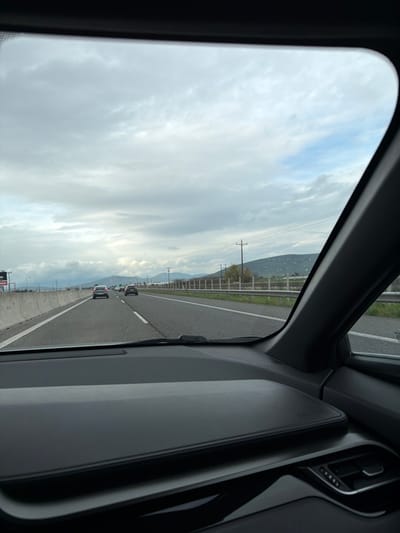 The image, captured from the interior of a vehicle looking through the windshield, depicts a multi-lane highway within the Municipal Unit of Thebes, Greece, most likely the A1 motorway (E75). The foreground shows a dark gray automotive dashboard, with the upper portion of the windshield framing the view ahead.
The highway consists of at least two visible lanes for traffic moving away from the viewer, marked by white dashed and solid lines. Ahead, three passenger vehicles, predominantly light-colored sedans or hatchbacks, are observed traveling in the same direction. To the left of the roadway, a concrete barrier extends along the highway. To the right, a grassy shoulder is bordered by a metal guardrail, beyond which lie green vegetation, several wooden utility poles with overhead power lines, and distant rolling hills or mountains.
The sky is overcast with a mix of gray and lighter clouds, revealing patches of blue sky in the upper right. No individuals are visible outside the vehicles. The scene portrays vehicular movement on a major transportation artery through a rural landscape under cloudy conditions.