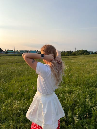 In this vertical, outdoor shot taken in Kutlu-Bukash, Russia, a person with long, blonde hair is seen from the back, standing in a grassy field. They are wearing a white t-shirt and a white skirt, with a hint of red fabric visible at the hem of the skirt. The person's hands are raised, with one hand touching their hair and the other near their head, suggesting they might be adjusting their hair or posing. A smartwatch is visible on their left wrist. A tattoo of a branch with leaves is on their left forearm.

The setting is a natural landscape with tall green grass and scattered wildflowers, illuminated by the soft light of either sunrise or sunset, casting a warm glow. In the distance, a line of trees borders the field, and behind them, there are some buildings and power lines, indicating a rural or semi-rural environment. The sky is a clear, pale blue, with a faint wispy cloud or contrail. The overall mood is serene and peaceful, capturing a moment of quiet reflection or enjoyment of nature. The time of day appears to be golden hour, given the warm lighting.